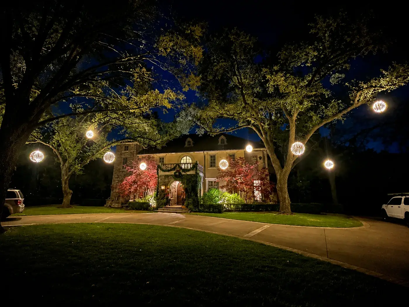 Glowing light orbs hanging from oak trees framing a grand DFW home with garland and wreath at the entry